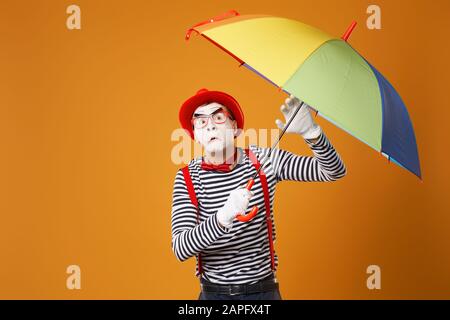 Trauriger MIME mit Blick auf die Kamera mit mehrfarbigem Regenschirm in der Hand isoliert auf leerem orangefarbenem Hintergrund im Studio Stockfoto