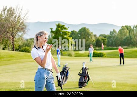 Frau spricht auf dem Smartphone auf dem Golfplatz, Freunde spielen im Hintergrund Golf Stockfoto