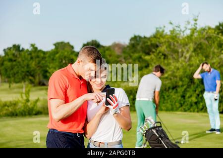 Paare, die Smartphone auf dem Golfplatz nutzen, Freunde, die im Hintergrund Golf spielen Stockfoto