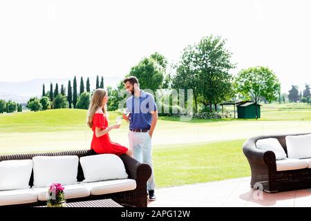 Ein Paar mit Weingläsern, die neben dem Sofa auf dem Golfplatz sprechen Stockfoto