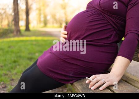 Nahaufnahme der schwangeren Frau mit rauchender Zigarette in der Hand, auf der Bank im Park sitzend. Geringe Schärfentiefe mit Fokus auf Zigarette. Stockfoto