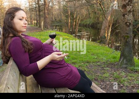 Schwangere Frau mit Alkohol und Zigarette in den Händen, die auf der Bank im Park sitzt. Seitliches Hochformat mit Kopierbereich Stockfoto