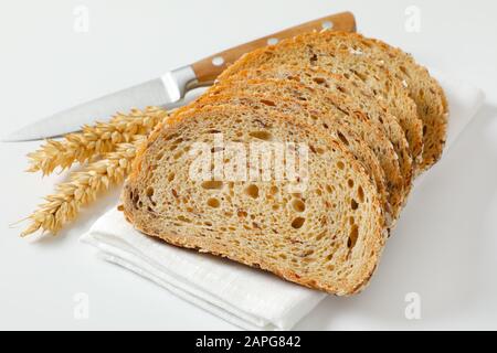 Brotlaib aus Vollkornbrot auf weißer Serviette, Weizenähren und Küchenmesser daneben Stockfoto