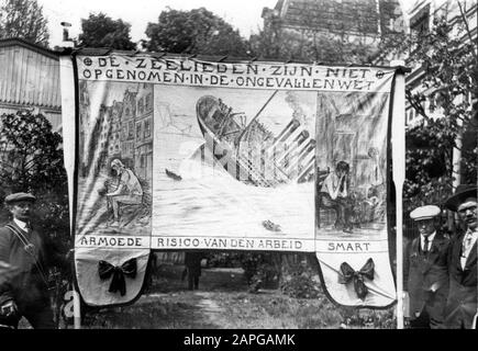 Banner der Seeleute in der Parade am 1. Mai 1914, Amsterdam. Sleuze an der Spitze: "Die Seeleute sind nicht im Unfallgesetz enthalten". Unter drei Bildern mit Bildunterschrift. Resp: Ein Seemann traf den Bettler ("Poverty"), eine überlebende Seemannswitwe mit Kind in der Wiege ("Smart") und einen sinkenden Ozeandampfer ähnlich der Titanic ("Risiko der Arbeit"). Amsterdam, Niederlande 1. Mai 1914. Stockfoto