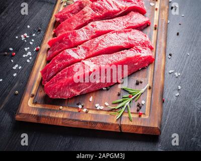 Rohes Fleisch, Rindersteak mit Würze auf Schneidebrett auf dunklem Hintergrund mit Rosmarin, Stockfoto