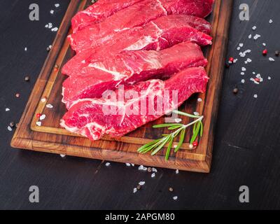 Rohes Fleisch, Rindersteak mit Würze auf Schneidebrett auf dunklem Hintergrund mit Rosmarin, Stockfoto