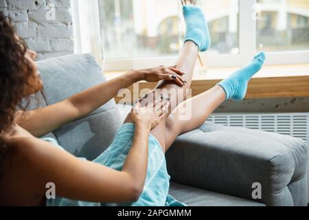 Beauty Day. Nahaufnahme der Frau im Handtuch, die ihren täglichen Hautpflegevorgang zu Hause macht. Auf dem Sofa sitzen und die Haut des Beines mit Feuchtigkeitscreme übersäen. Konzept von Schönheit, Selbstpflege, Kosmetik, Jugend. Stockfoto