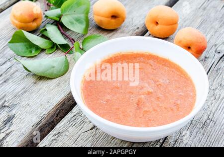 Weiße Schüssel mit hausgemachter Aprikosenmarmelade auf einer Holzoberfläche in der Nähe reifer Aprikosenpfanne und grüner Blätter. Nahaufnahme. Stockfoto
