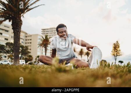 Lächelndes Porträt eines jungen Sportlers, der auf grünem Gras sitzt und Dehnübungen im Park macht Stockfoto