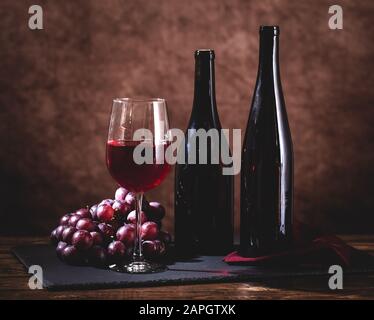 Glas Rotwein mit Trauben und zwei Weinflaschen auf einem Holztisch Stockfoto