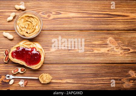 Sandwich mit Erdnussbutter anstoßen. Löffel und Glas Erdnussbutter, Marmelade und Erdnüsse zum Kochen des Frühstücks auf braunem Holzgrund. Cremige Erdnuss Stockfoto