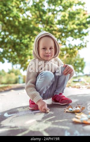 Kleiner Junge 3-5 Jahre alt, glücklich zeichnet bunte Flusskrebse auf dem Straßenbelag, Kreativität im Park, fühlen sich glücklich. Legere warme Kleidung mit Kapuze. Herbsttag Stockfoto