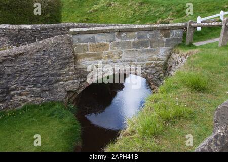 Eine sehr kleine Bogenbrücke aus Backsteinen und Stein über einen schmalen blauen Bach, der zwischen hohen grasigen Ufern fließt. Malerisches und niedliches Beispiel für das E Stockfoto