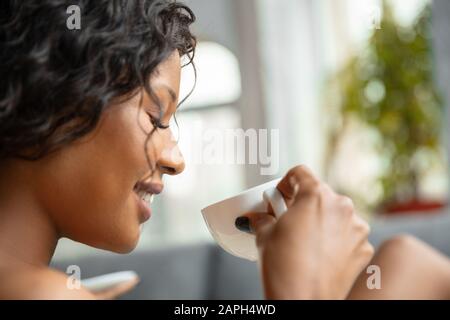 Nahaufnahme der afroamerikanischen Frau in Handtuch, die ihren täglichen Schönheitsalltag zuhause macht. Auf dem Sofa zu sitzen, sieht zufrieden aus, trinkt Kaffee und entspannt. Konzept von Schönheit, Selbstpflege, Kosmetik, Jugend. Stockfoto