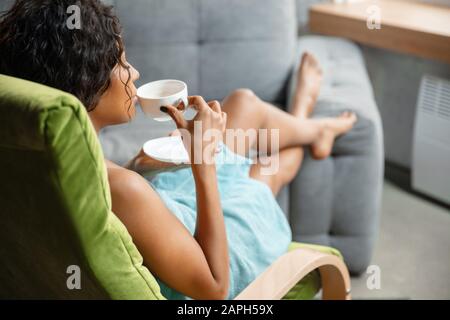 Chill Day. Afro-amerikanische Frau im Handtuch, die ihren täglichen Schönheitsalltag zuhause macht. Auf dem Sofa zu sitzen, sieht zufrieden aus, trinkt Kaffee und entspannt. Konzept von Schönheit, Selbstpflege, Kosmetik, Jugend. Stockfoto