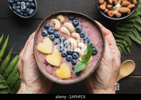 Tropische Bananen und Blaubeere Smoothie in den Händen der Frau. Gesundes Frühstück in der Kokosschale. Ansicht von oben. Sauberes Essen, vegetarisches Essen. Stockfoto