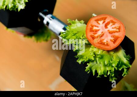 Eine Scheibe frische Tomate und Salat liegen auf einer großen Hanteln. Symbol für schwere Lebensmittel unter dem Deckmantel von frischem Gemüse. Schwere Lebensmittel Stockfoto