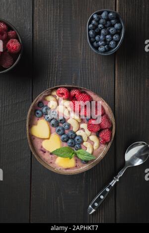 Bananen- und Blaubeer-Smoothie-verzierte Früchte in Kokosschale auf Holzbrett. Ansicht von oben. Sauberes Essen, vegetarisches Essen. Vertikales Format. Stockfoto