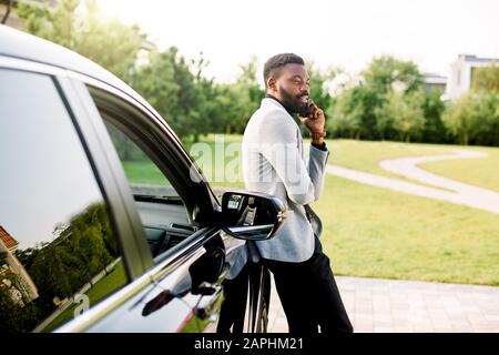 afroamerikanischer Geschäftsmann in einem Anzug, der auf dem Smartphone spricht, während er draußen in der Nähe seines luxuriösen schwarzen Autos steht. Grünpark, Bäume im Hintergrund. Stockfoto