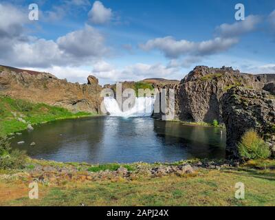 Epischer Luftdronblick, der an einem sonnigen Tag über eine Landschaft mit Wasserfall und Lagune von Hjalparfoss fliegt. Stockfoto