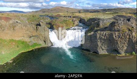 Epischer Luftdronblick, der an einem sonnigen Tag über die Landschaft des Hjalparfoss Wasserfalls und der Lagune fliegt.