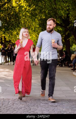 Lächelnde Hüftpaare essen Eis und haben Spaß in der Stadt: Stilvoller junger Mann mit Bart und blonde Frau in rotem Kleid laufen im Str Stockfoto