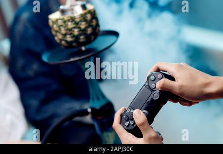 Mann hält Gamepad in der Hand, im Hintergrund Rauch von Wasserpfeifen. Stockfoto