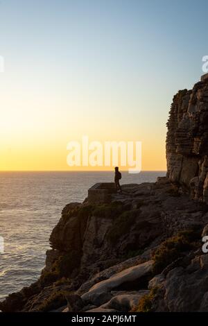 Eine Person in der Ferne, die am Rande einer Klippe steht und bei Sonnenuntergang auf den Ozean blickt. Ansicht von der Seite. Stockfoto