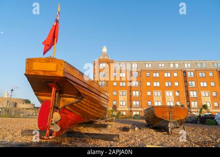 Sternblick auf zwei klassische, altmodische Boote, die am steinigen Strand hochgezogen wurden, mit einem großen roten Ziegel-Apartmentgebäude im Hintergrund, das in Licht der Sonne leuchtet Stockfoto