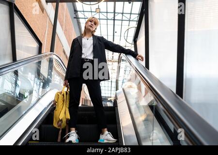 Blonde Haarfrau in halbformaler Kleidung, die mit breiten Beinen auf einer Rolltreppe steht, die den gelben Rucksack hält, und ihren Arm auf dem Handlauf aufhält, niedriger Winkelschuss. Stockfoto