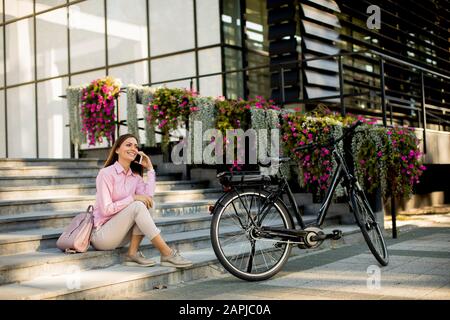 Junge Frau, die auf der Treppe in der Nähe von Ebike sitzt und Smartphone im Freien aushustert Stockfoto