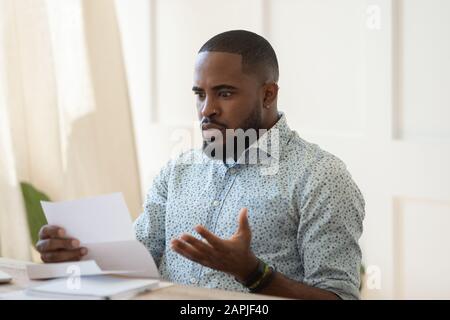 Afrikanischer Mann, der am Tisch sitzt und Brief liest, der schreckliche Nachrichten liest Stockfoto