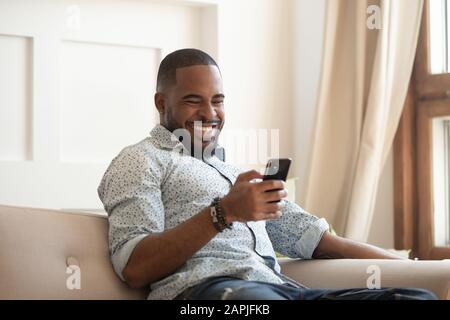 Fröhlicher afrikanischer Kerl, der auf der Couch sitzt und Zeit mit dem Smartphone verbringt Stockfoto