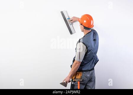 Die Builder glättet die Wand mit einem Spachtel. In Arbeitskleidung und harten Hut. Auf einem hellgrauen Hintergrund. Stockfoto