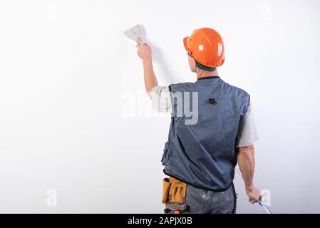 Die Builder glättet die Wand mit einem Spachtel. In Arbeitskleidung und harten Hut. Auf einem hellgrauen Hintergrund. Stockfoto