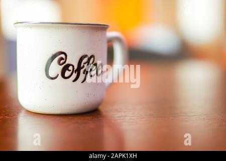 Emaille weiße Metallbecher mit Aufschrift Kaffee auf Holztisch auf verschwommenem Hintergrund. Platz für Text oder Werbung Stockfoto