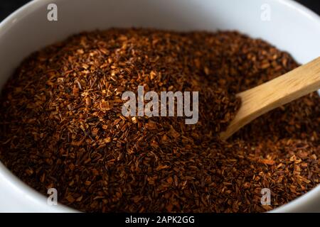 Nahaufnahme von getrockneten Rooibos (Redbush) Teeblättern in einer weißen Keramikschale mit Holzlöffel. Stockfoto