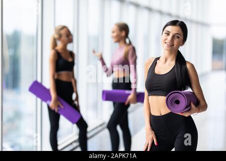 Junge Fitness-Frau hält Matte vor zwei fit-Freundinnen, die im Fitnessstudio stehen Stockfoto