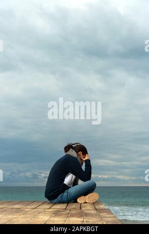 Frau sitzt mit dem Kopf in den Händen am Meer, launischer Himmel, Depression, Konzept der psychischen Gesundheit. Stockfoto
