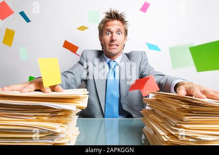 Gestresster Geschäftsmann, der auf den Schreibtisch hält, um zwischen großen Papierstapeln zu sitzen Stockfoto