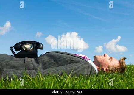 Entspannter Geschäftsmann, der auf einem sonnigen Flecken von hellgrünem Gras schläft, mit einem altmodischen Drehtelefon, das auf seinem Anzug ruht Stockfoto