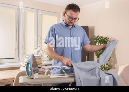 Mann im Wohnzimmer Bügeleisen die Ärmel eines Hemdes auf einem Bügelbrett. Im Hintergrund liegen gebügelte und gefaltete Handtücher auf der TA Stockfoto