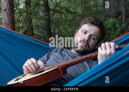 Nachdenklicher bärtiger Mann liegt in einer Hängematte mit einer akustischen Gitarre in den Händen und spielt eine Melodie gegen einen Wald. Stockfoto