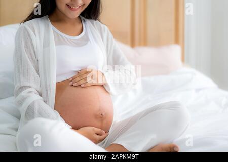 Nahaufnahme der lächelnden, schwangeren asiatischen Frau, die auf dem Bett sitzt und zu Hause ihren Bauch berührt. Schwangerschafts-, Ruhe-, Menschen- und Erwartungskonzept Stockfoto