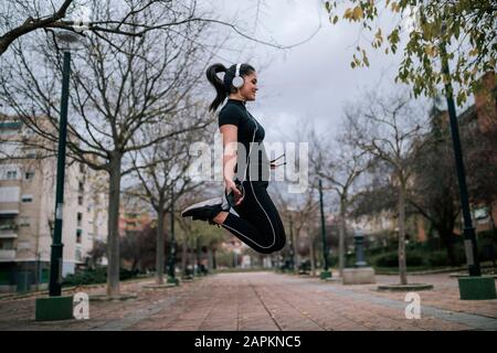 Junge Frau in schwarzer Sportswear springt in die Luft und hört Musik mit Kopfhörern Stockfoto