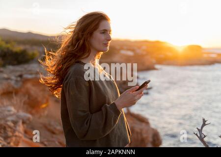 Redheaded junge Frau mit Handy an der Küste bei Sonnenuntergang, Ibiza, Spanien Stockfoto