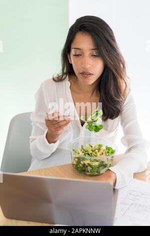 Porträt des jungen Architekten, der gemischten Salat am Schreibtisch mit Blick auf den Laptop isst Stockfoto