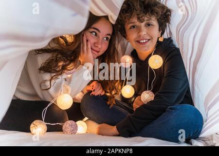 Porträt eines glücklichen Bruders und einer Schwester mit einer Kette von Lichtern unter dem Bettbezug Stockfoto