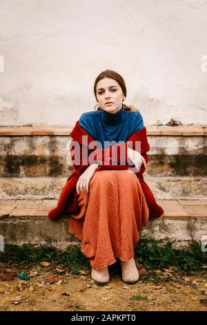 Nahaufnahme des Frauenporträts mit blauem Turtleneck-Pullover und rotem Mantel auf einer Stufe sitzend Stockfoto