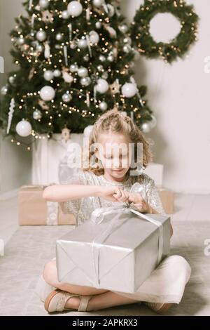 Porträt des blonden kleinen Mädchens, das vor dem Weihnachtsbaum sitzt, das Weihnachten öffnet Stockfoto